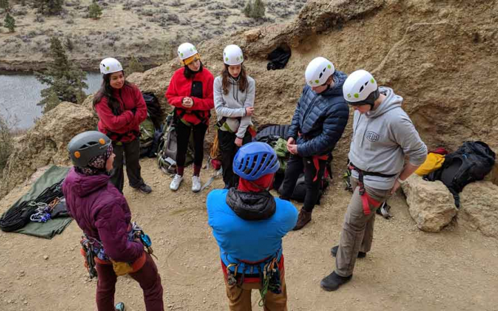 a group of adults watch an outward bound instructor tie a knot on a rock climbing course